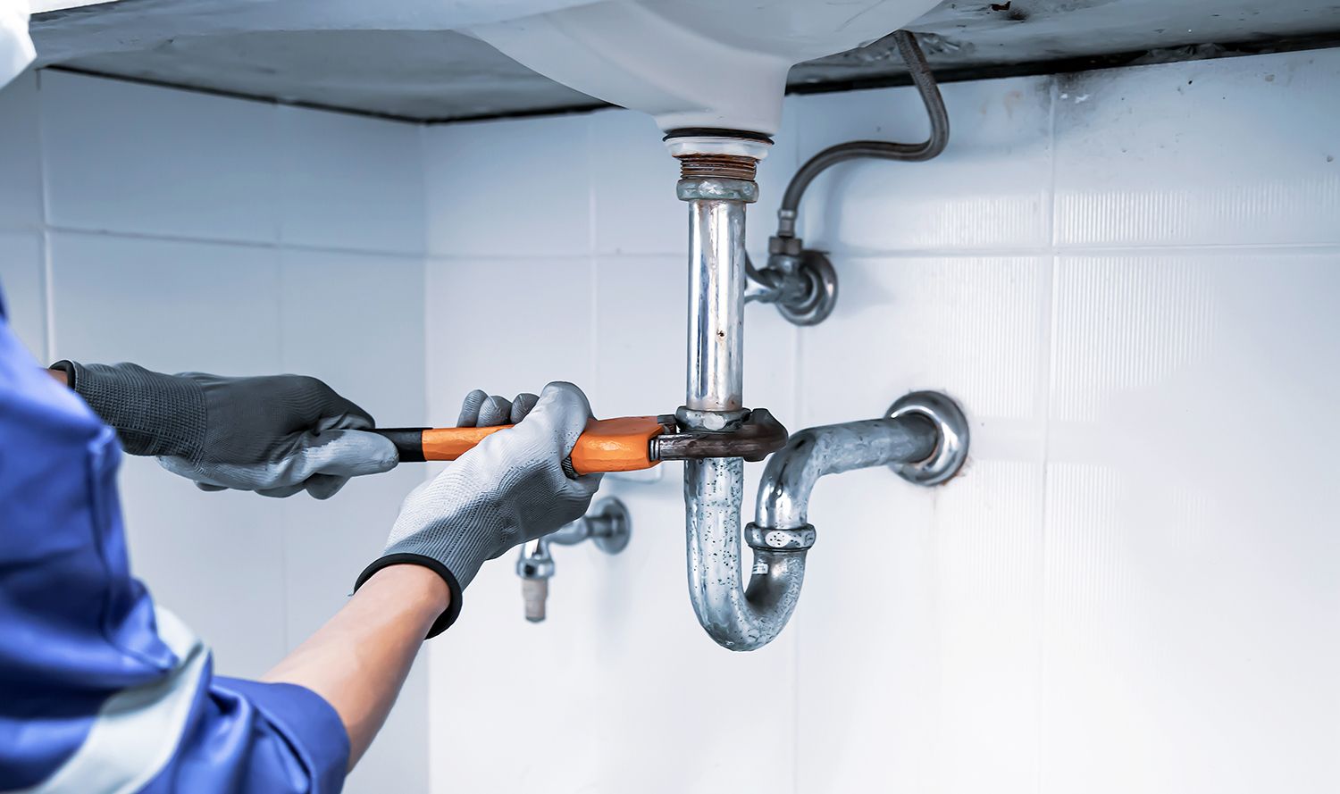Plumber in blue uniform uses a wrench to tighten a pipe under a white sink.