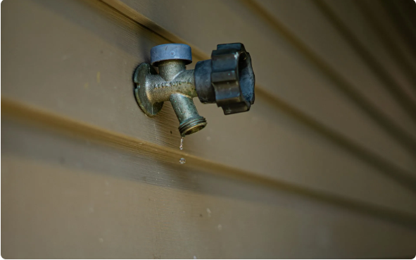 A brass outdoor water spigot mounted on brown horizontal siding, with a black plastic handle and a small leak.