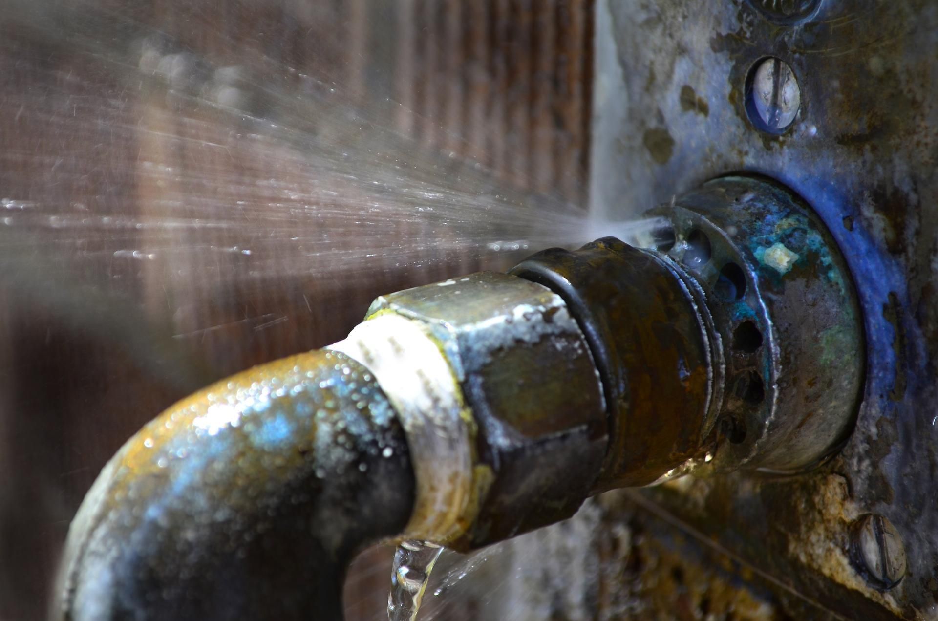 A close-up of a metal pipe connection leaking a fine spray of water against a textured wall surface.