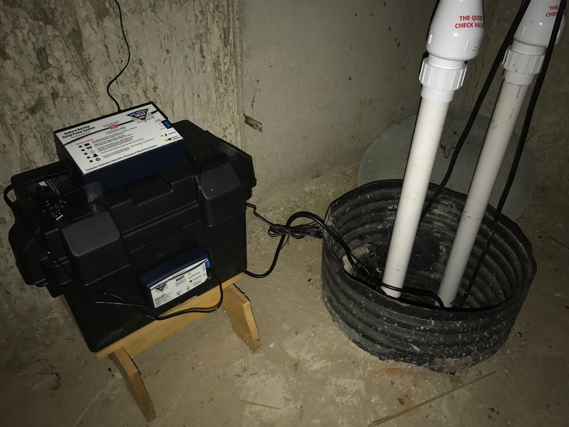 A sump pump system in a basement featuring a battery backup unit in a black case connected to a pump basin via PVC pipes.