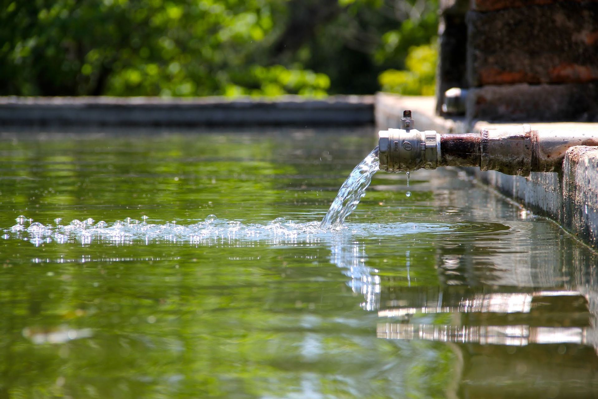 Water flows from a metal pipe into a green outdoor pool on a sunny day.