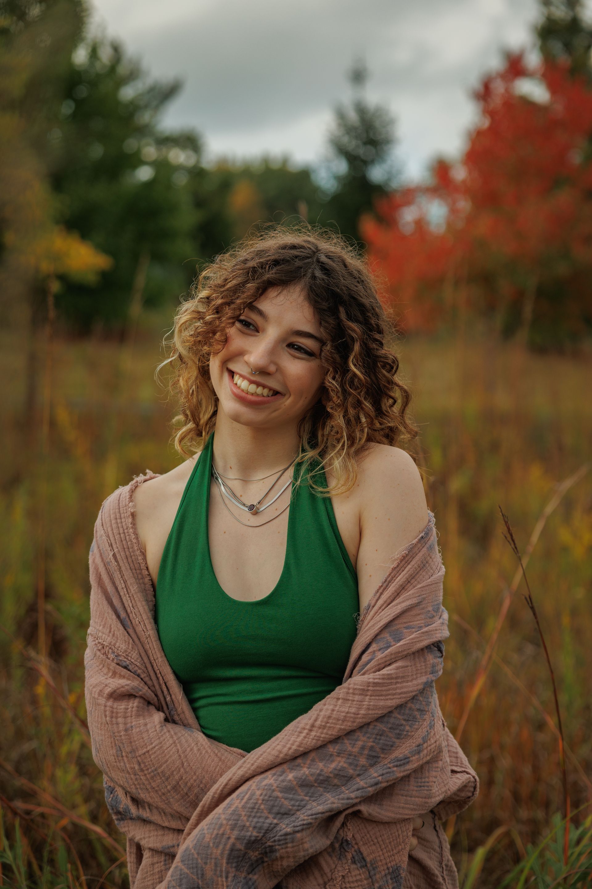 Young woman smiling in a garden, wearing a white top and black cardigan. She is near a flowering tree, surrounded by greenery.