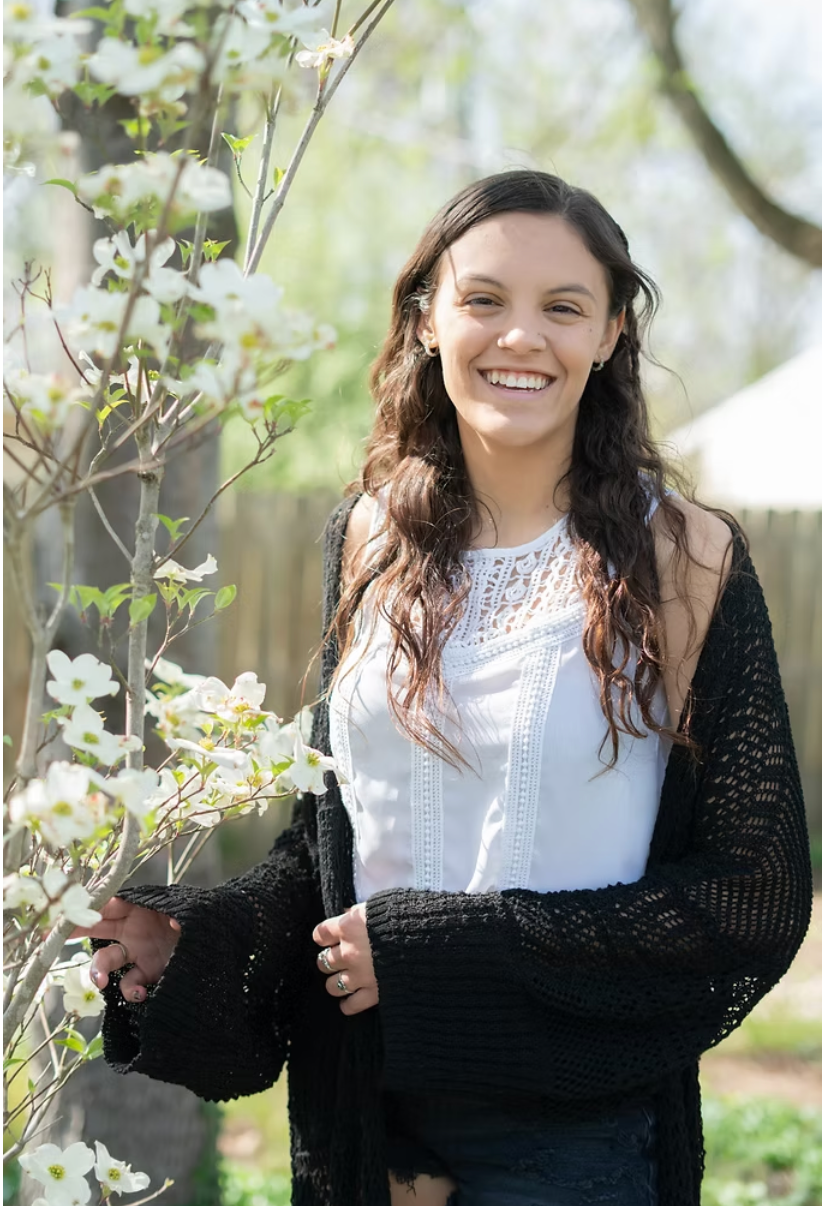 Young woman smiling in a garden, wearing a white top and black cardigan. She is near a flowering tree, surrounded by greenery.