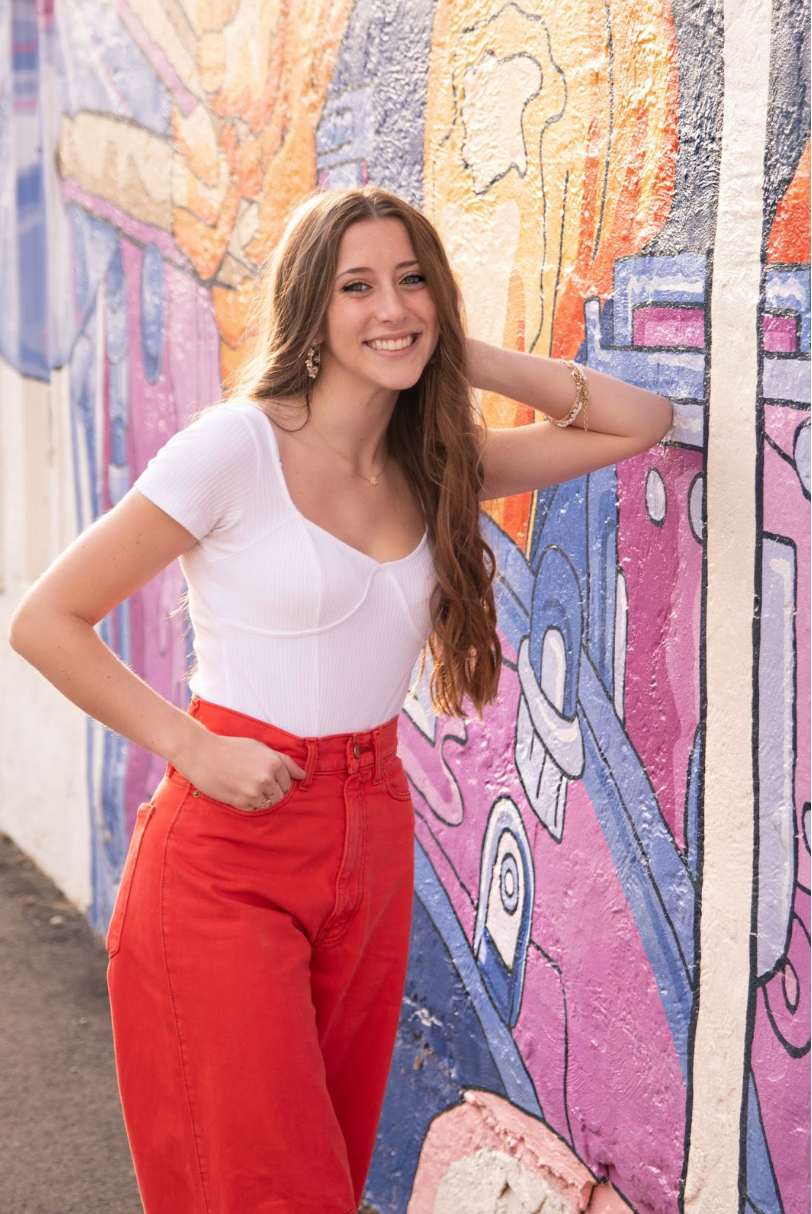 Young woman smiles, leaning against a colorful mural. She wears a white top and red pants.
