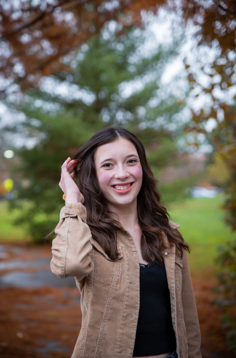 Smiling teen girl in a tan jacket and black top, touching her hair with a park backdrop in autumn.