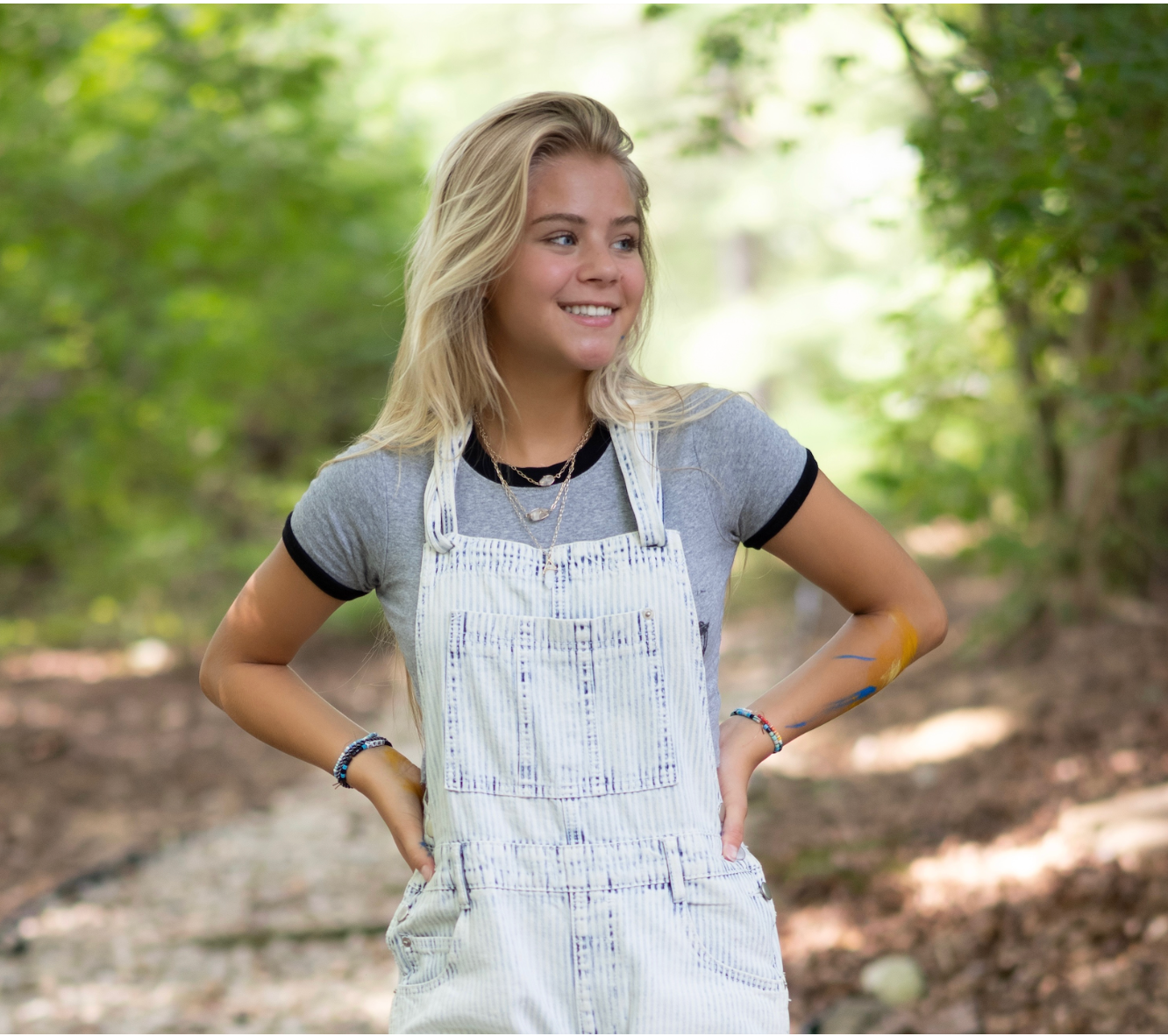 Blonde girl in overalls smiles, standing outdoors with hands on hips.