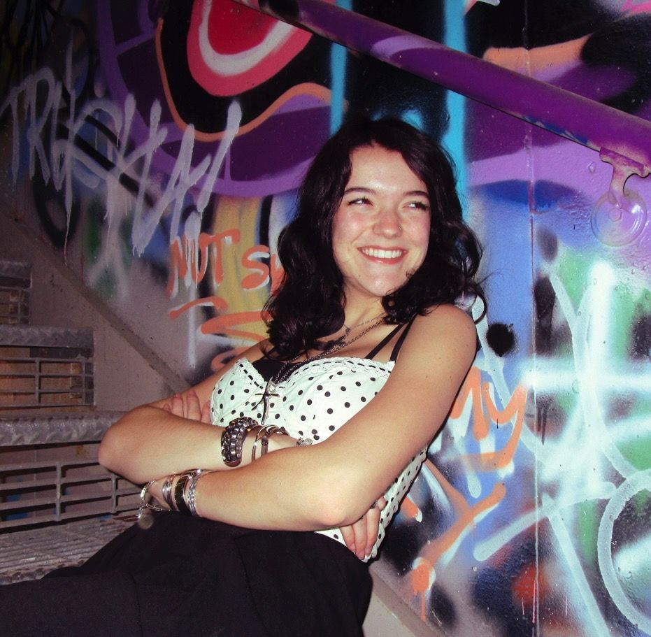 Young woman smiles, leaning against a colorful mural. She wears a white top and red pants.