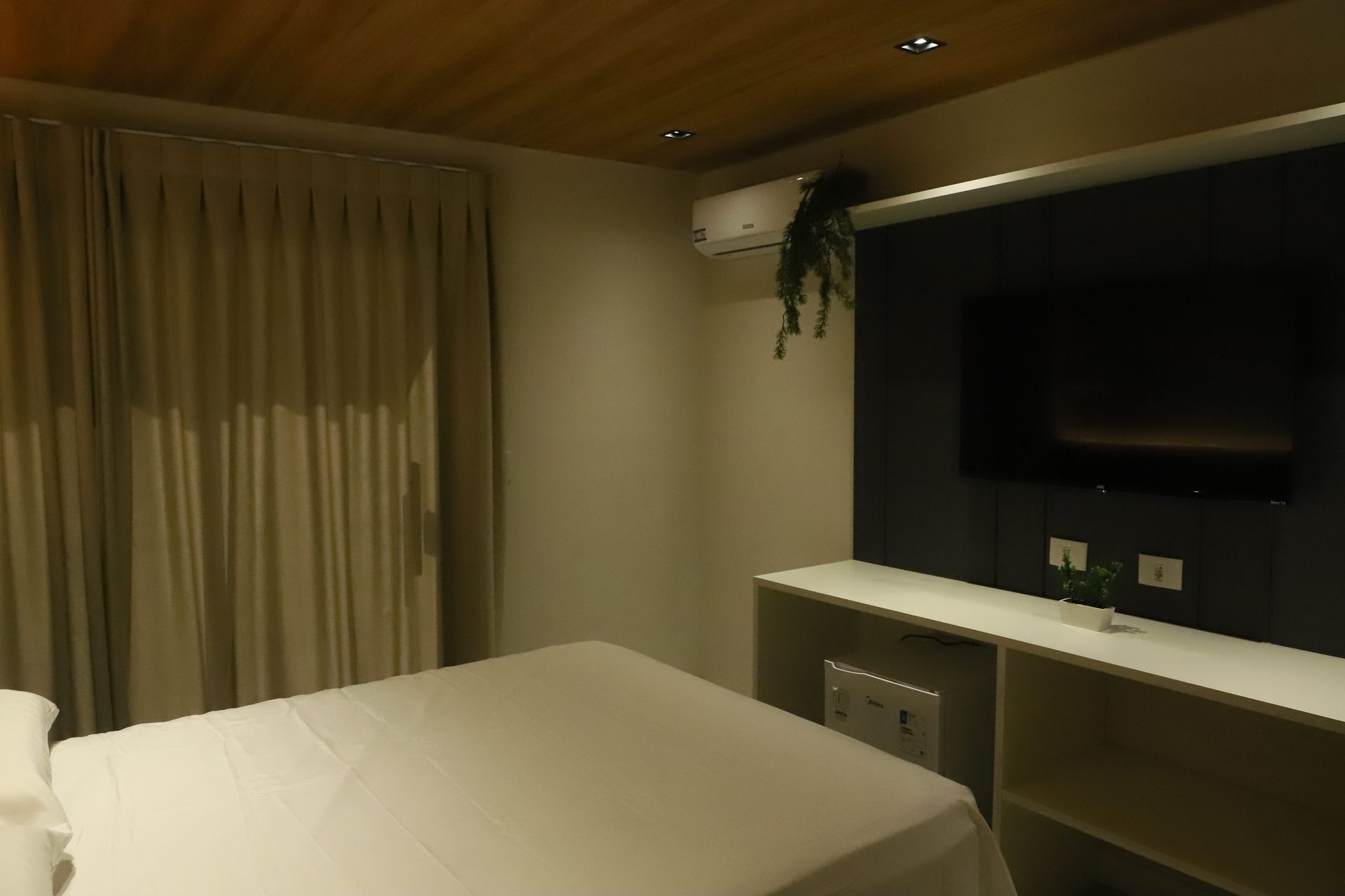 Quarto de hotel com pouca luz, cama arrumada, cortinas, TV de parede e prateleira com uma planta pendurada.