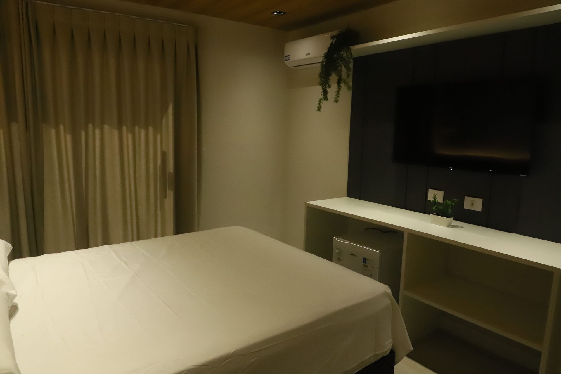 Quarto de hotel com cama, janela com cortina, TV de parede e escrivaninha branca.