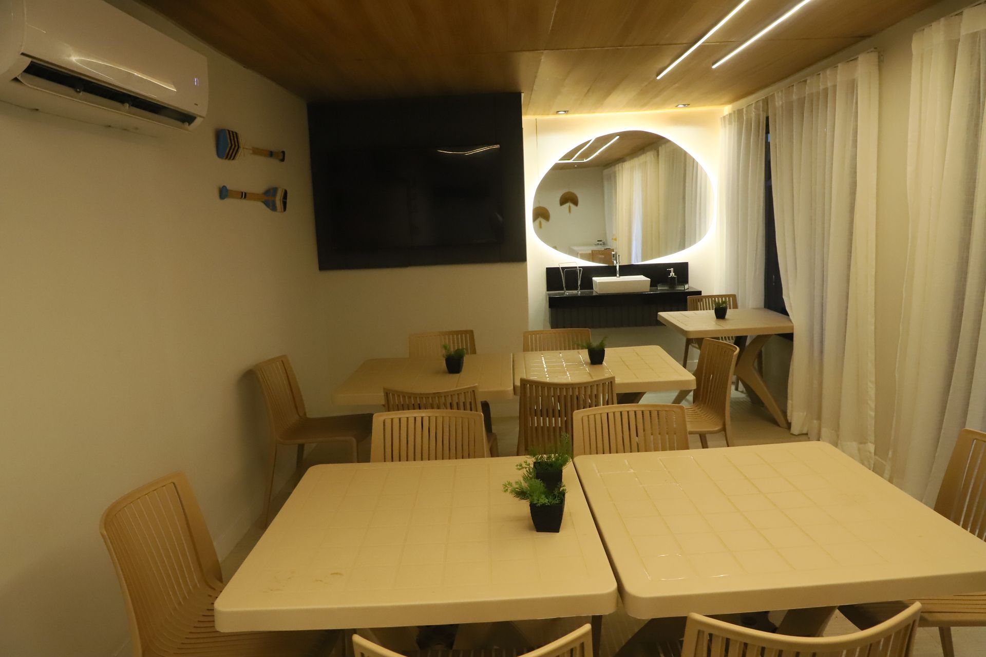 Área de jantar aconchegante com mesas, cadeiras de vime, TV fixada na parede e cortinas translúcidas.
