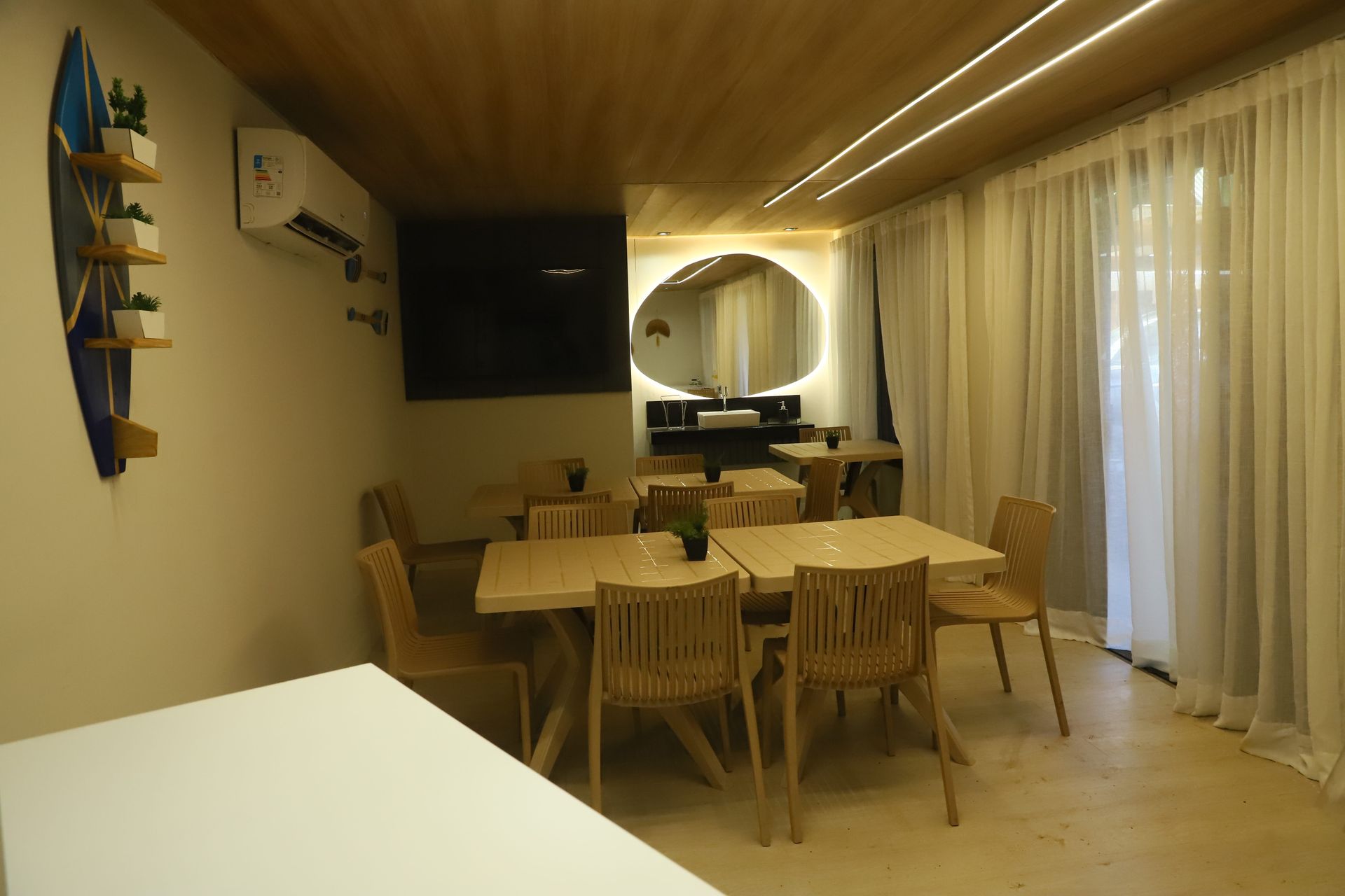 Sala de jantar moderna com mesa e cadeiras de madeira, TV de parede e cortinas translúcidas junto a uma janela bem iluminada.