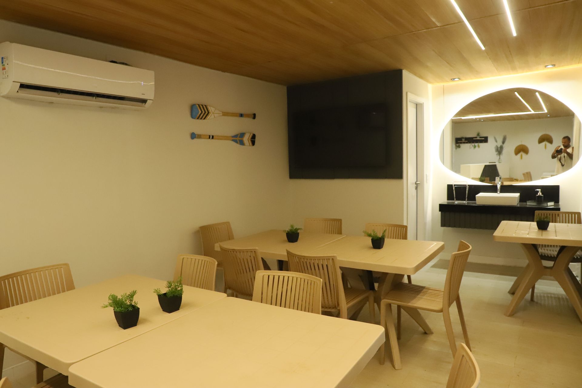 Interior minimalista de um café com mesas e cadeiras de madeira, obras de arte na parede, ar condicionado e uma TV fixada na parede.