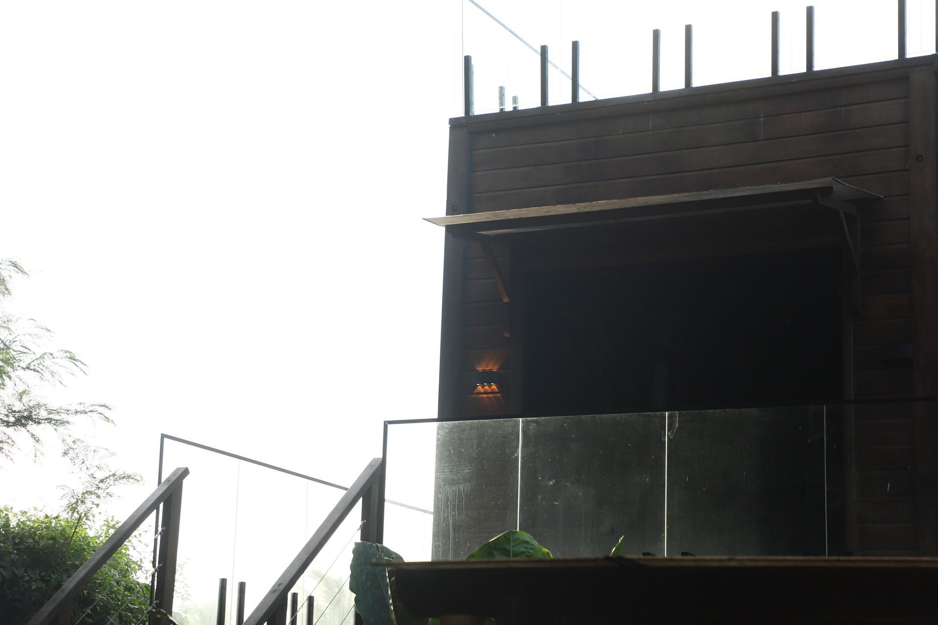 Estrutura de madeira escura com guarda-corpo de vidro e escadas à esquerda, sob luz solar intensa.