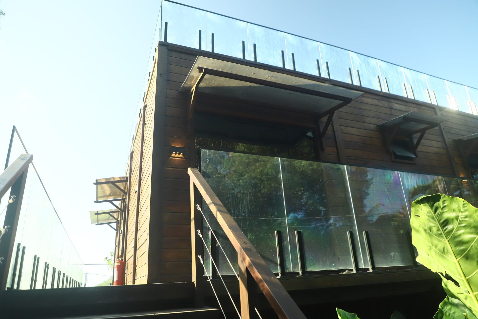 Exterior de casa moderna com varanda de vidro e revestimento de madeira sob luz solar intensa.