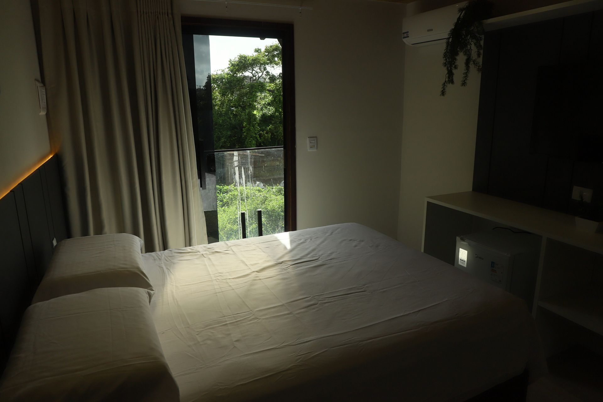 Quarto pouco iluminado com cama desarrumada, porta aberta para um pátio verde e TV à direita.