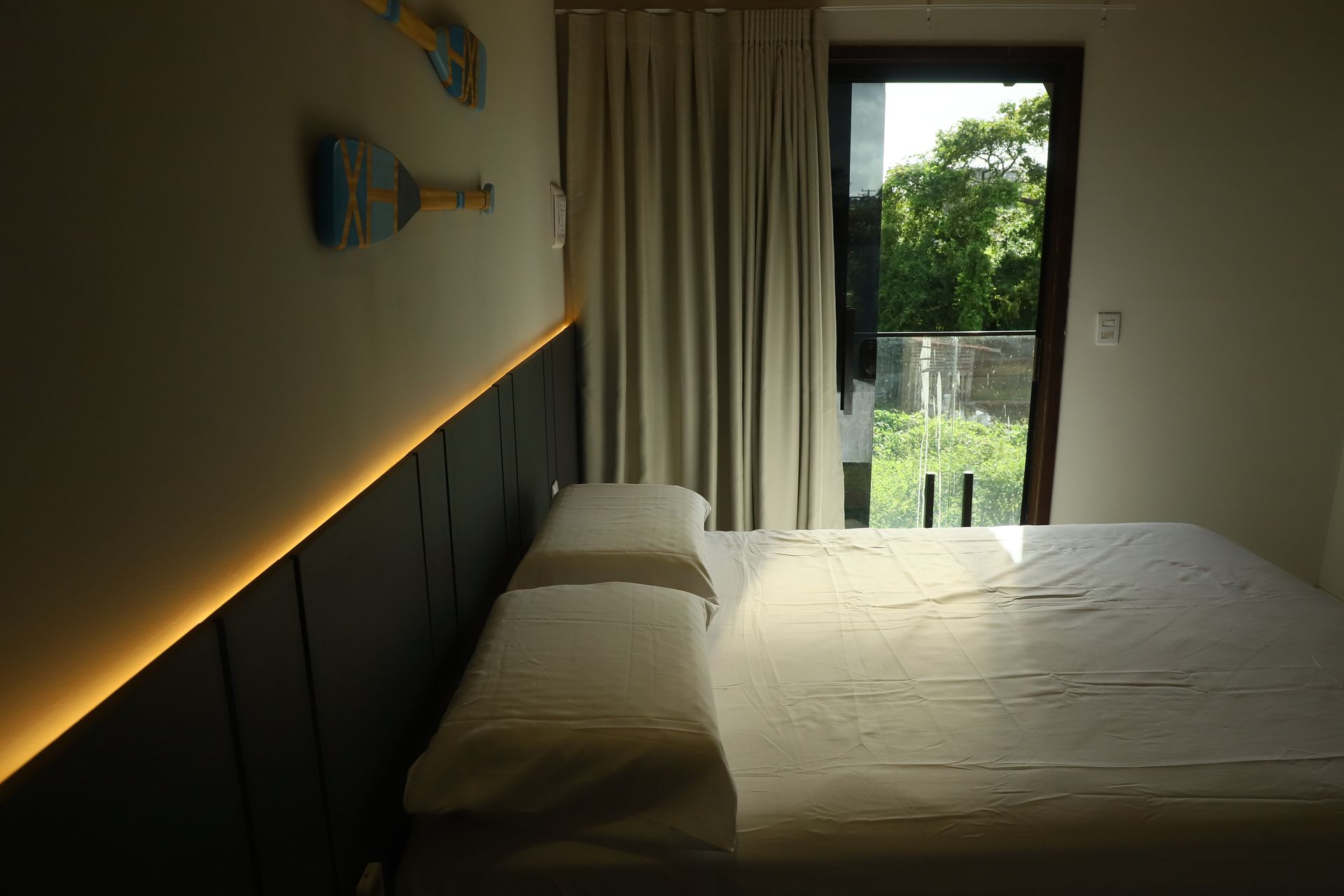 Quarto com cama, iluminação LED aconchegante na parede, cortinas e porta de varanda com vista para a vegetação.