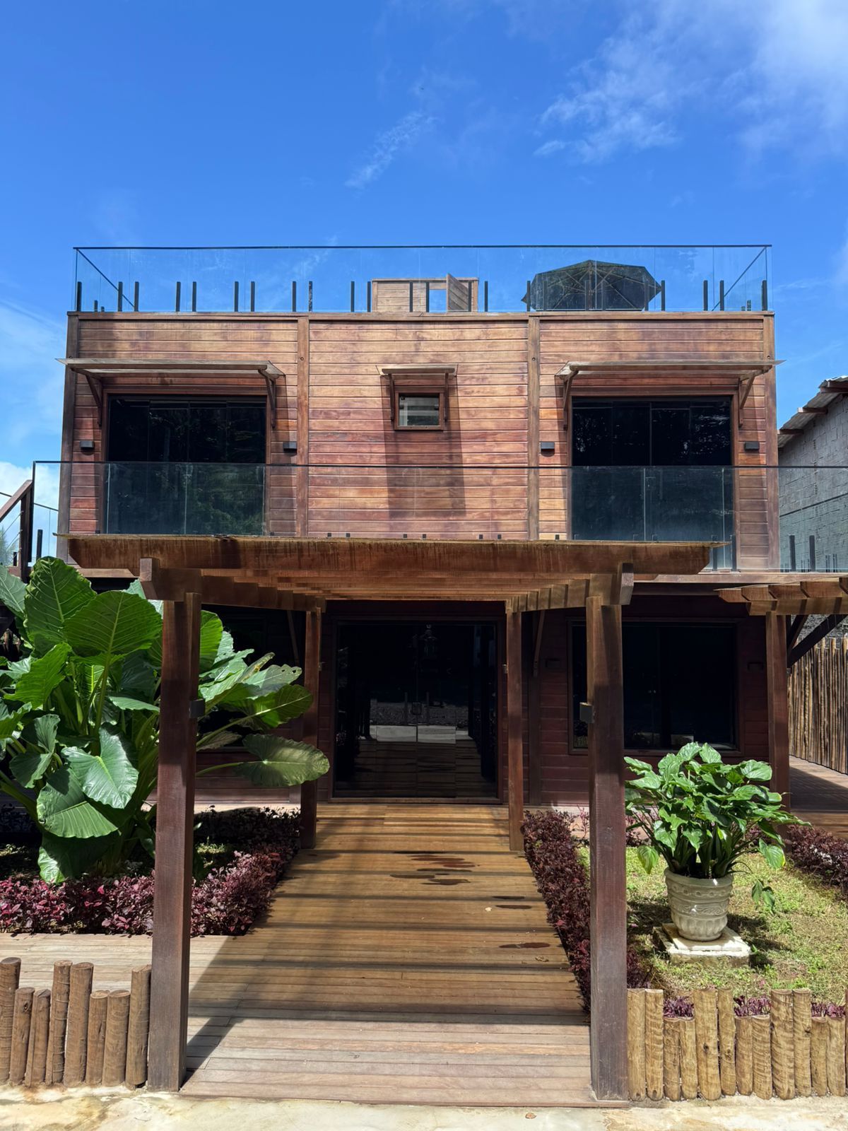 Casa de praia de madeira com deck frontal, escadas e plantas tropicais sob um céu azul límpido.