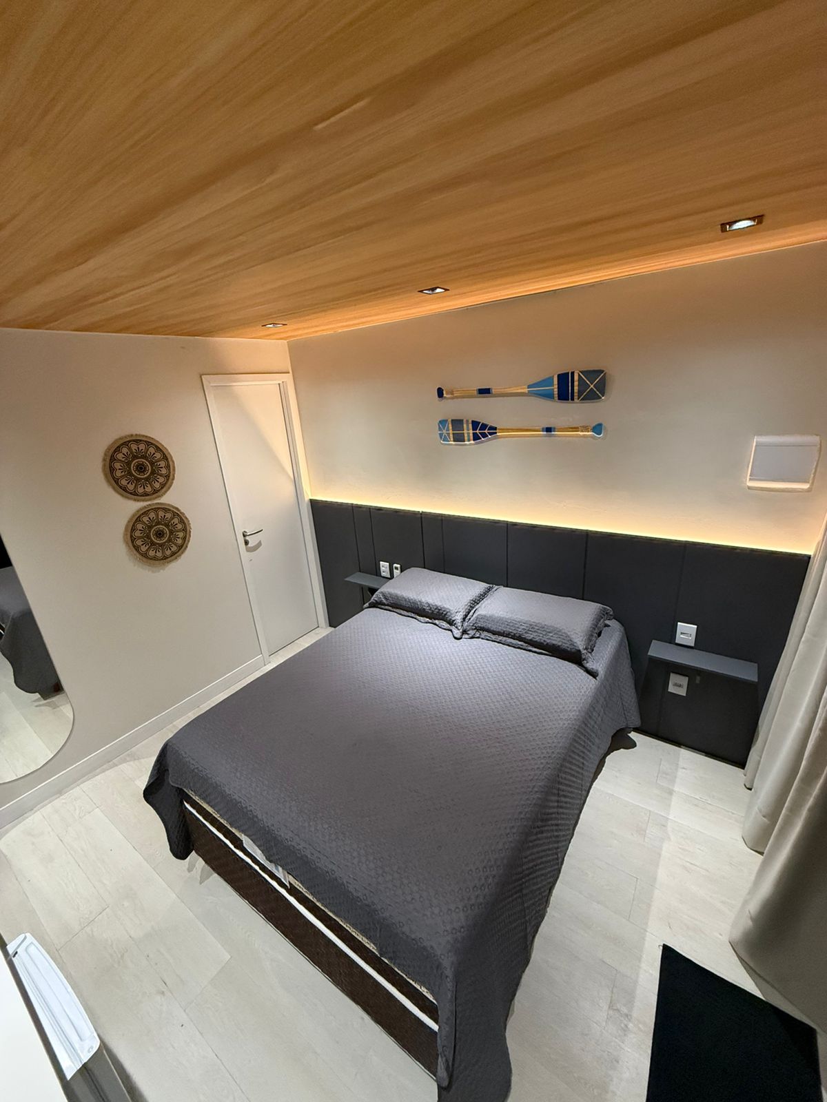 Quarto moderno com cama cinza, teto de madeira, paredes brancas e pranchas de surfe penduradas acima da cabeceira.