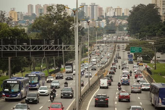 Rodovia movimentada de várias faixas com tráfego intenso, viaduto e prédios da cidade ao longe sob um céu enevoado.