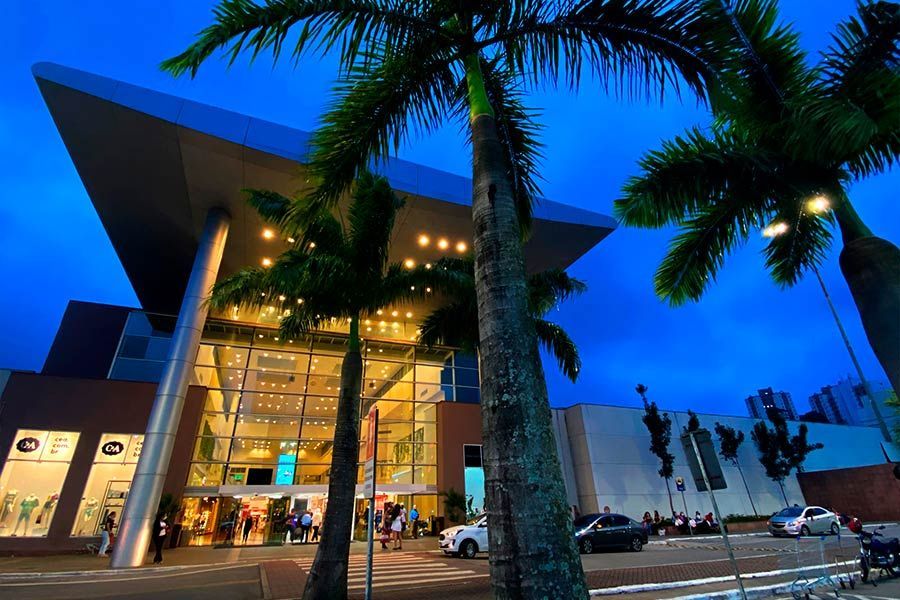 Edifício moderno com fachada de vidro ao entardecer, com palmeiras e iluminação interna aconchegante.