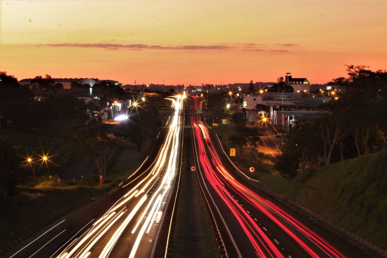 Longa exposição em rodovia ao pôr do sol com rastros de luzes brancas e vermelhas de carros sob um céu alaranjado.