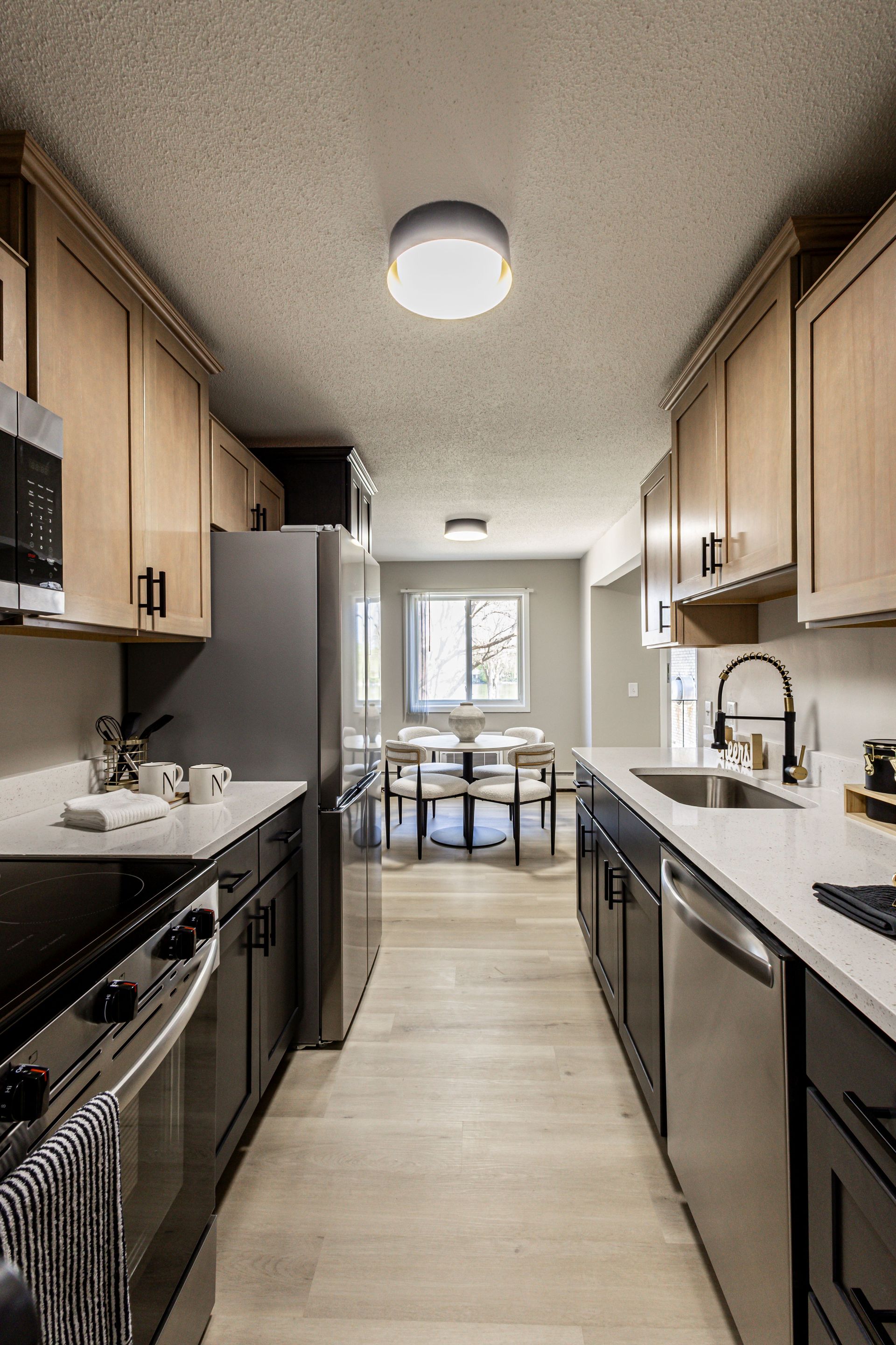 Modern galley kitchen with light wood cabinets, stainless appliances, and a dining area beyond