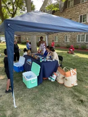 People at a backyard canopy booth with coolers, drinks, and bags on a grassy lawn.
