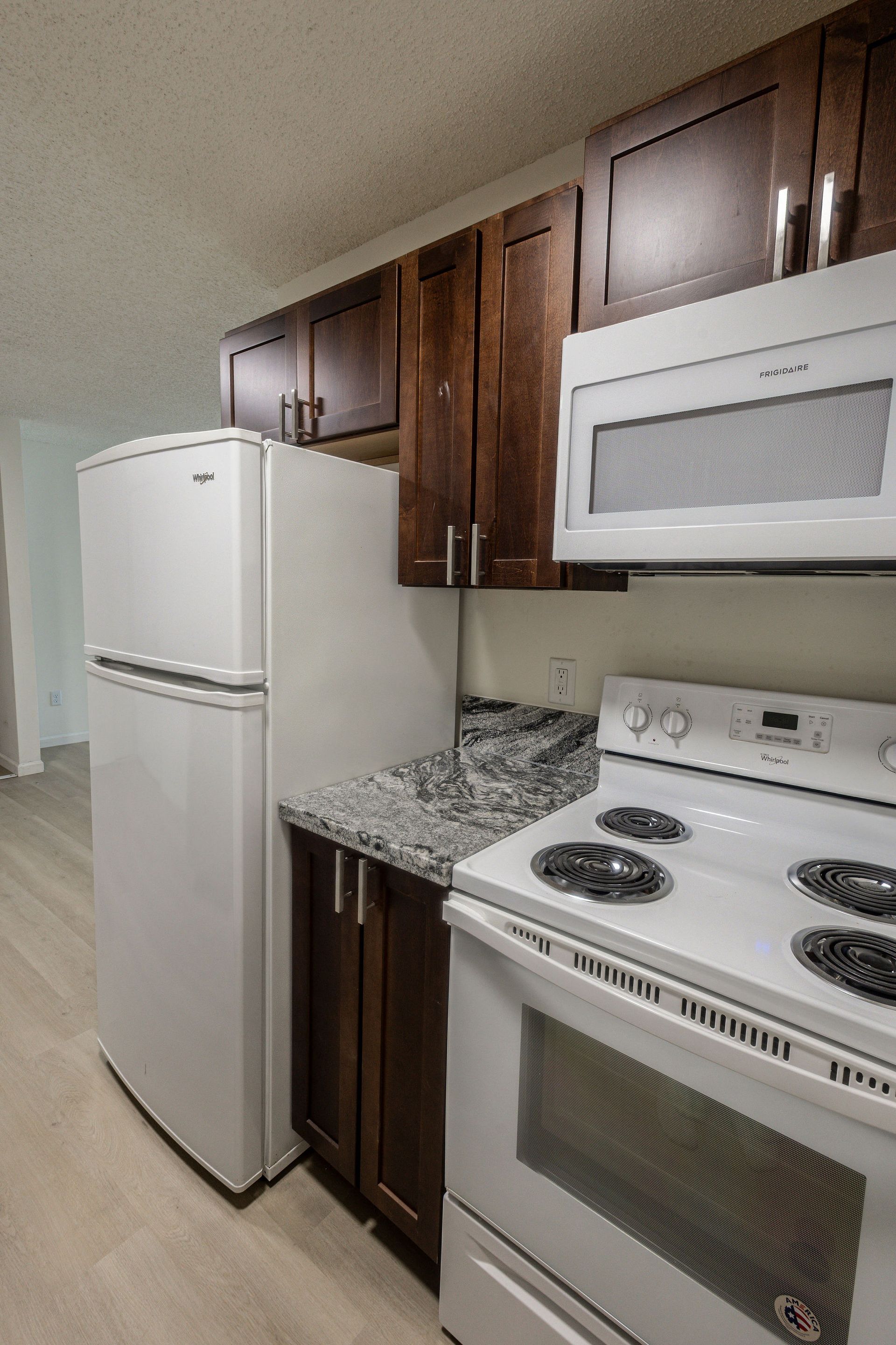Compact kitchen with white appliances, dark wood cabinets, and speckled countertops.