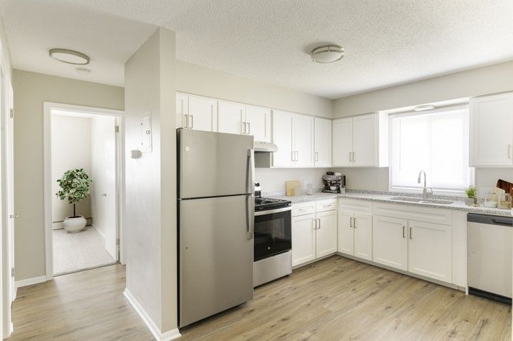 Bright white kitchen with stainless steel appliances, light wood floors, and a doorway to a hall with a potted plant.