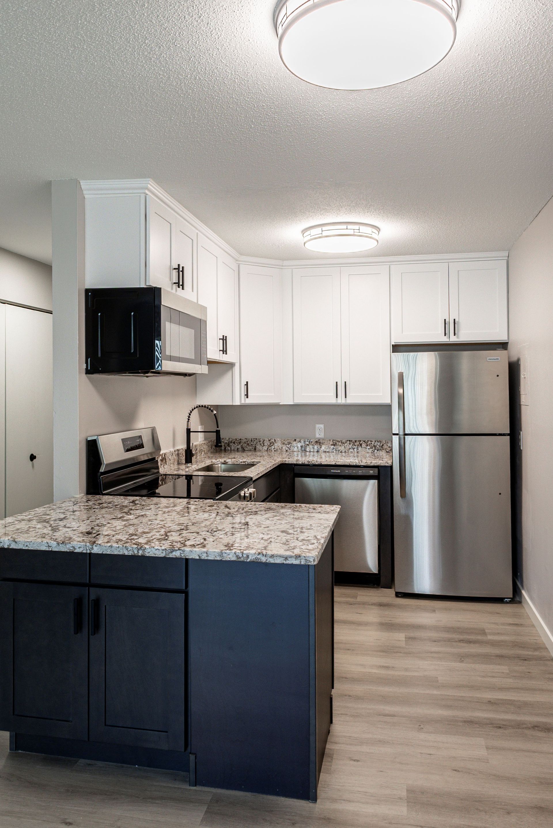 Modern kitchen with dark island cabinets, white upper cabinets, granite counters, stainless fridge, and wood floor