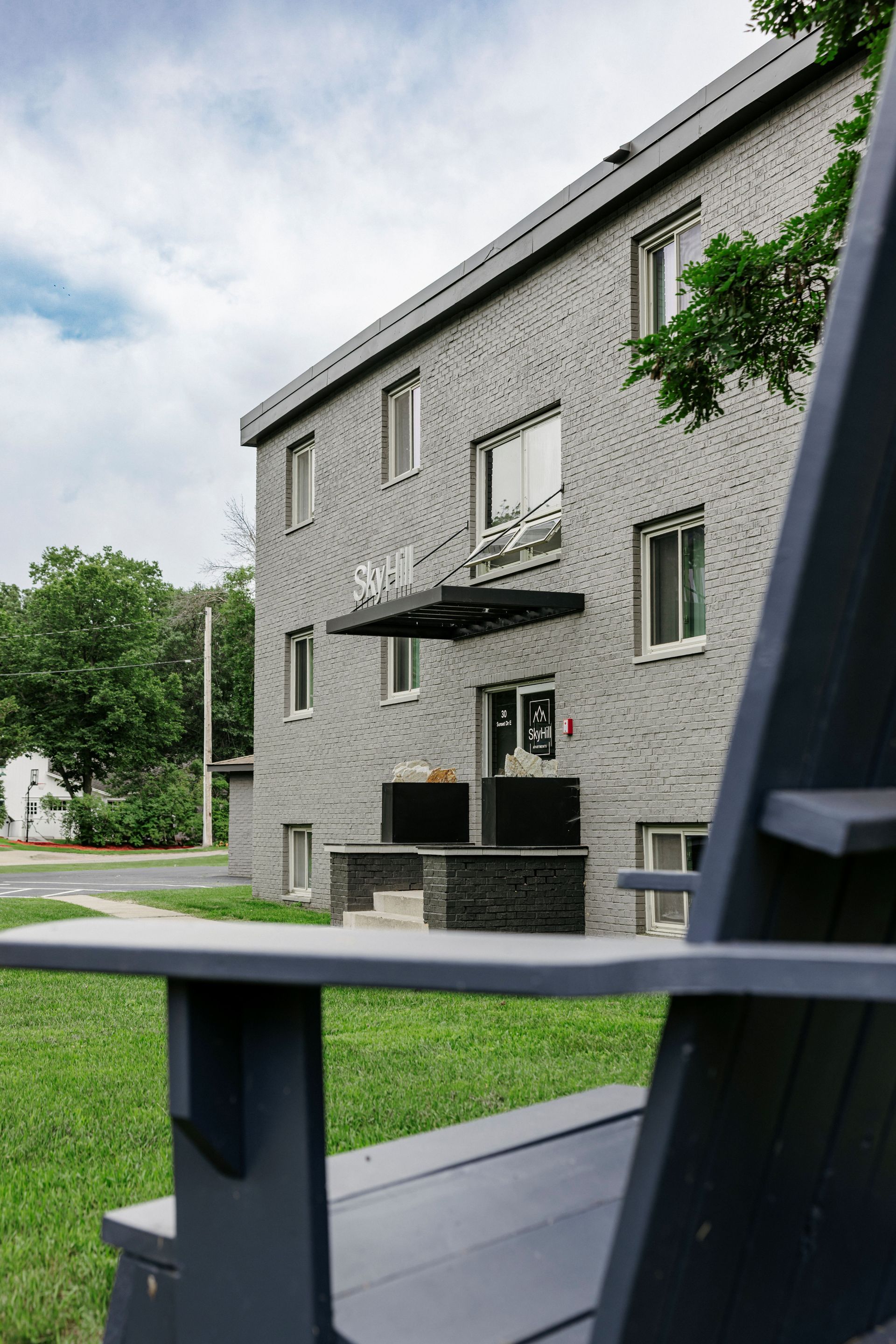 Gray apartment building with balconies beside a green lawn and patio chair in the foreground