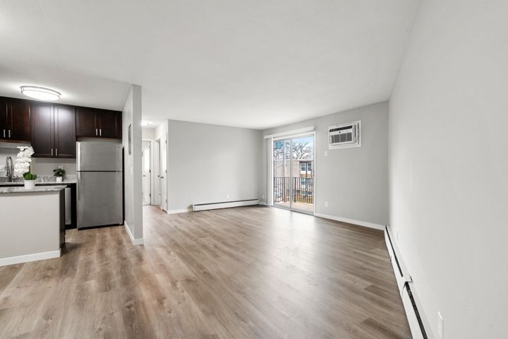 Empty modern apartment living room with wood floors, kitchen corner, sliding glass door, and wall AC unit
