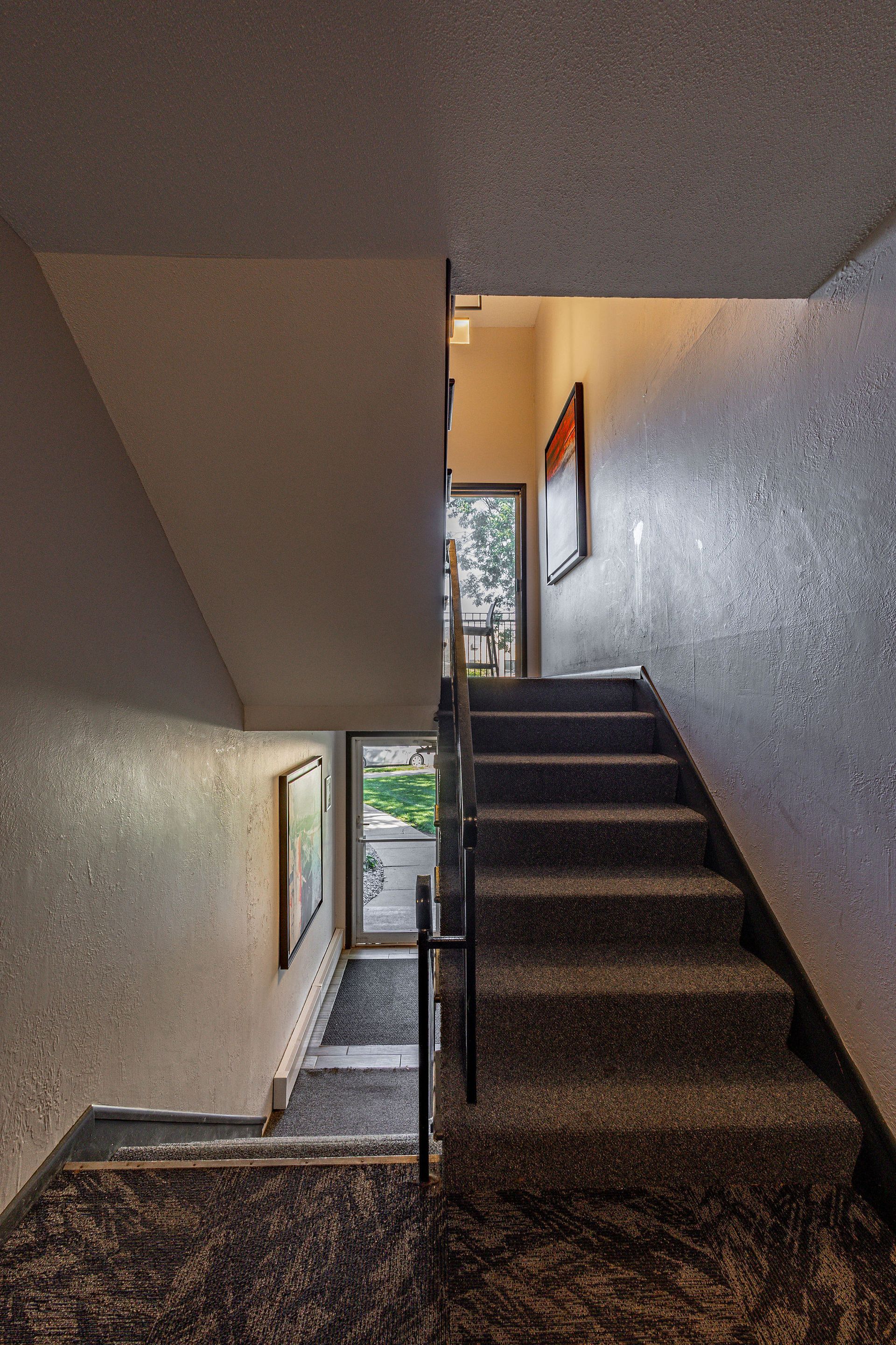 Narrow indoor stairwell with carpeted steps and white walls leading to a bright exit door below.