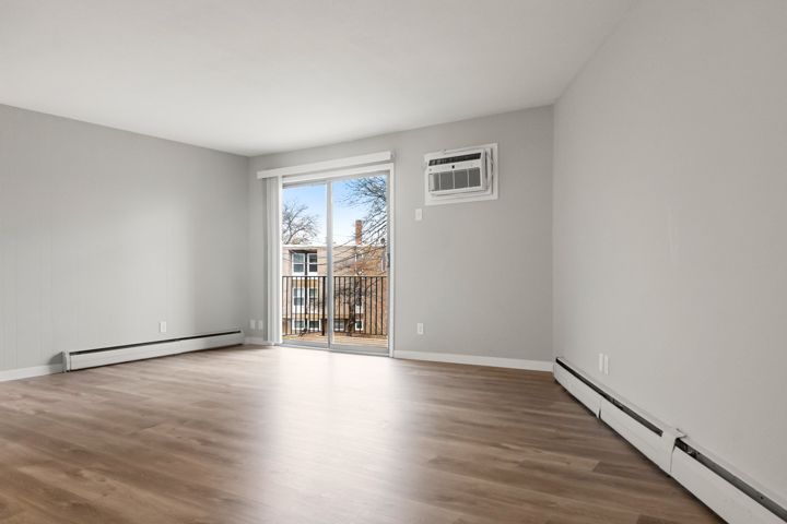 Empty apartment room with light wood floors, white walls, sliding glass door, and wall AC unit