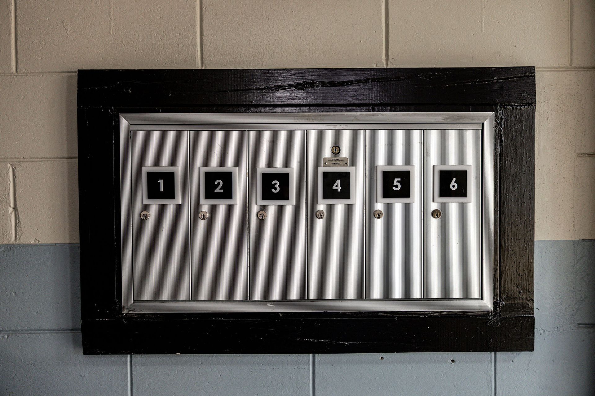 Six-unit wall-mounted mail slot panel with numbered compartments, black frame, and beige background.