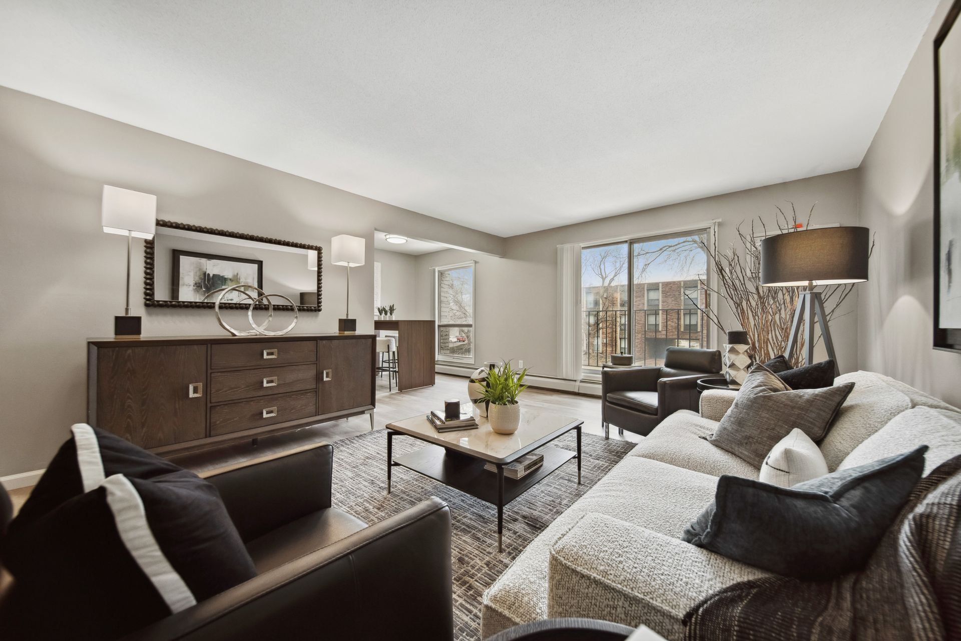 Bright living room with gray sofa, dark armchair, wood dresser, coffee table, and large windows with city view