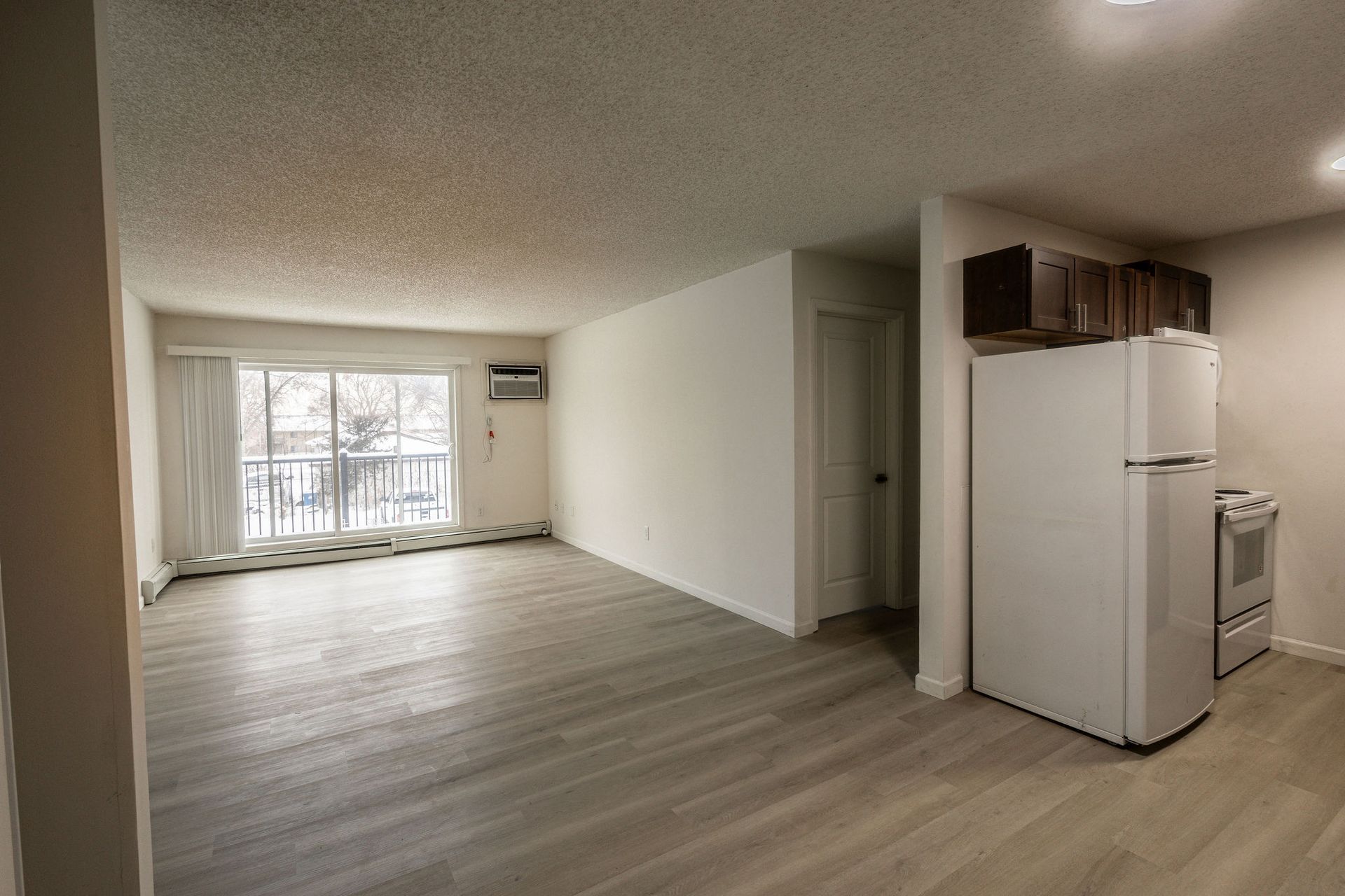Empty apartment living room with light wood floors, white walls, and a kitchen area on the right.