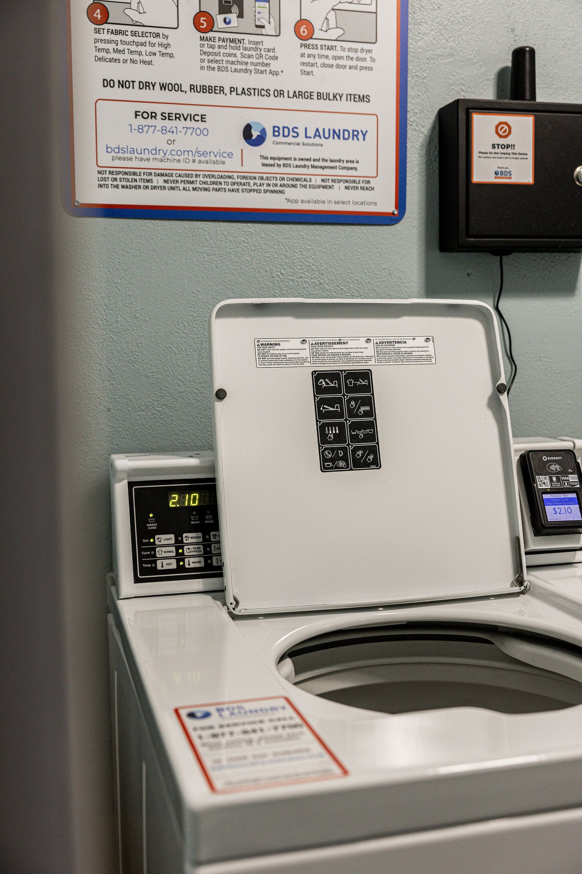 Laundry room with a top-loading washing machine, white lid open, next to wall signs and a payment box.