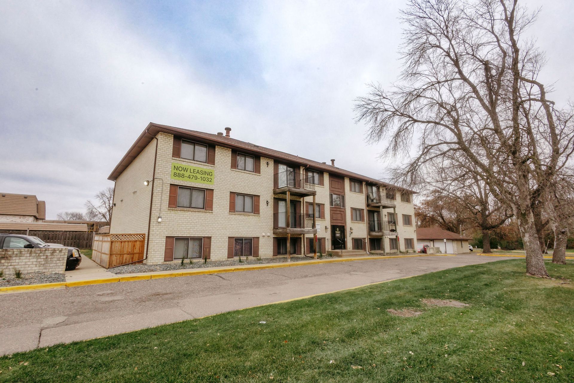 Apartment building with balconies beside a road and grassy lawn under a cloudy sky