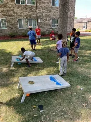Children playing cornhole on a grassy lawn beside a brick building, with beanbags and boards in view