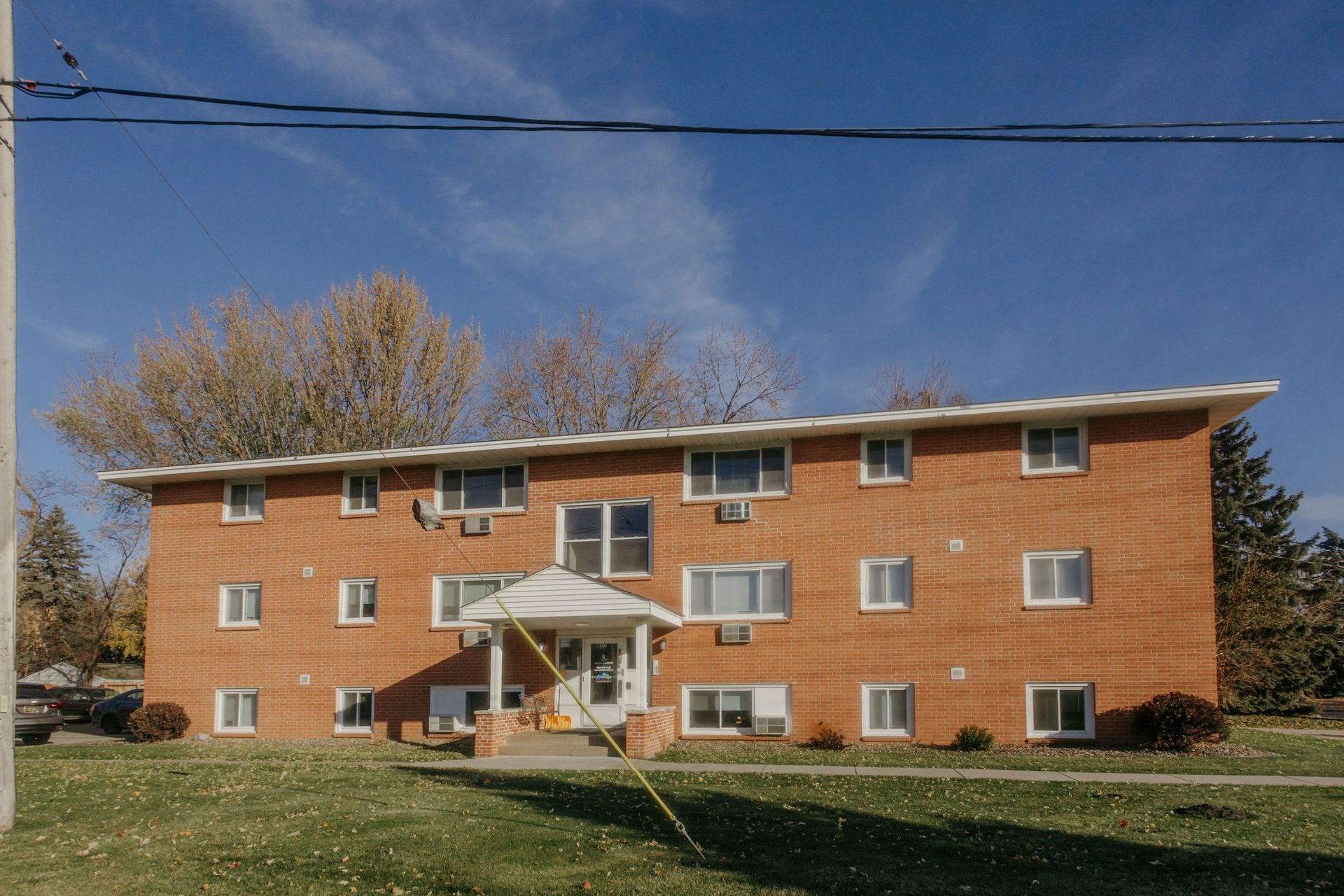 Brick apartment building with a white entrance porch and a grassy front lawn under a blue sky