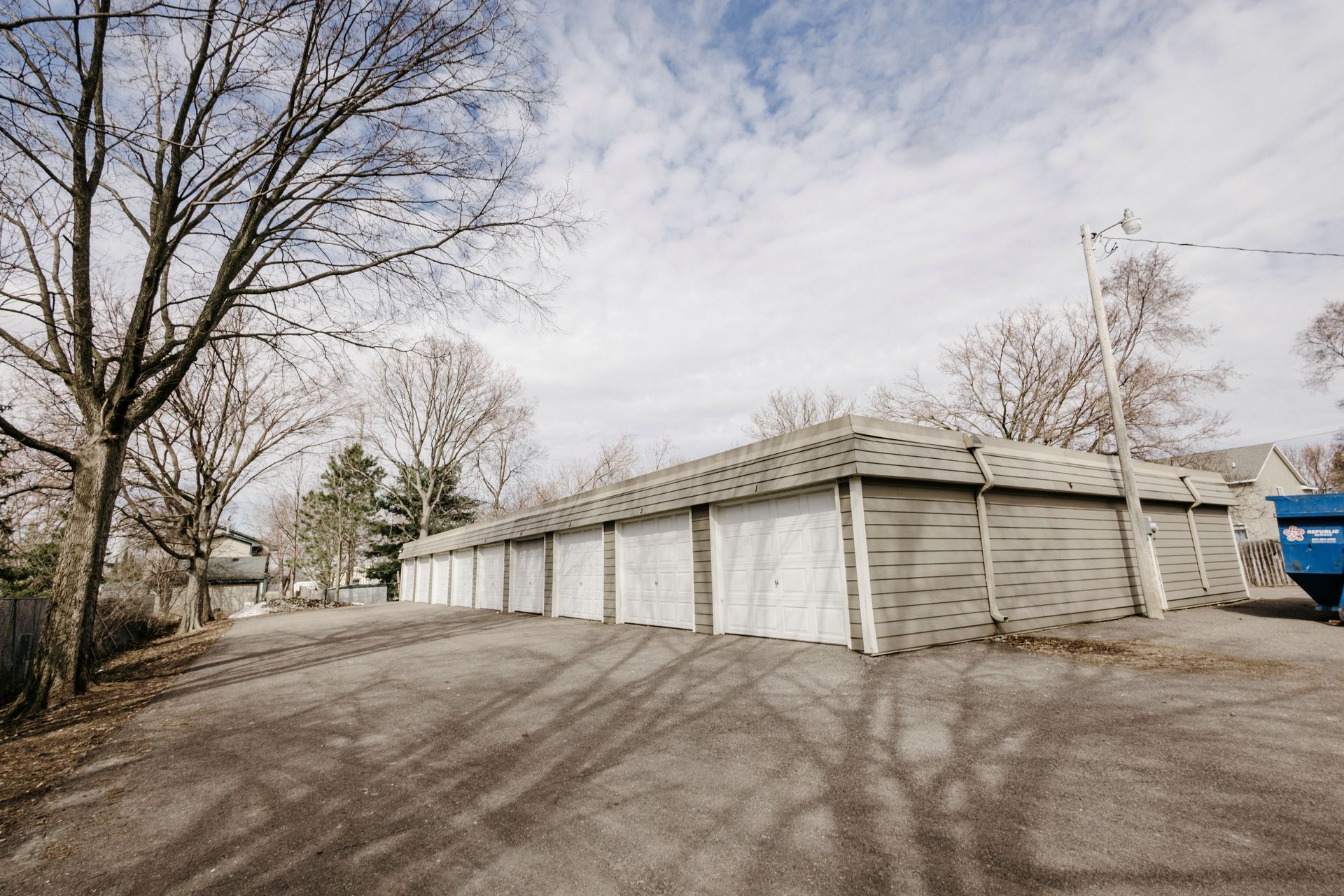 Row of white garage units beside a paved lot with leafless trees and an overcast sky