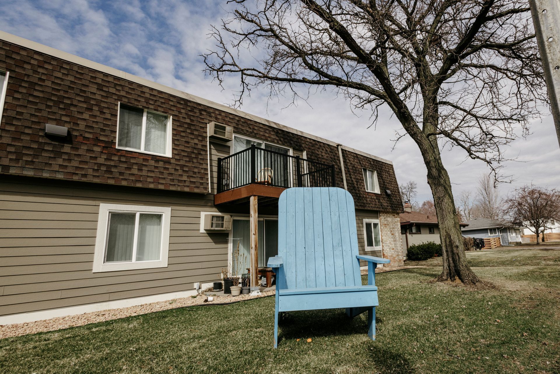 Blue Adirondack chair on a lawn in front of a beige apartment building with a tree overhead