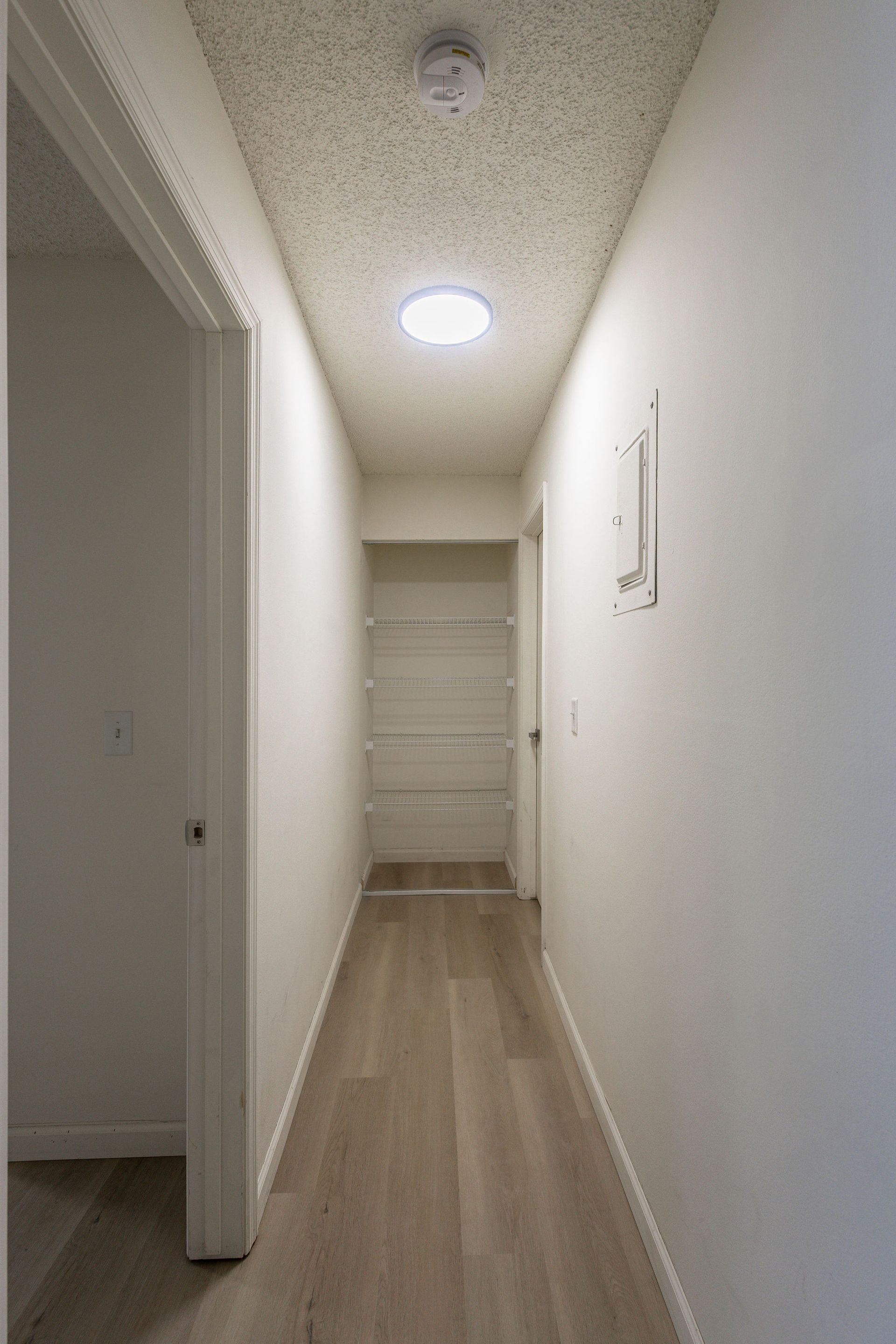 Narrow white hallway with wood floor, ceiling lights, and a closed door at the end.