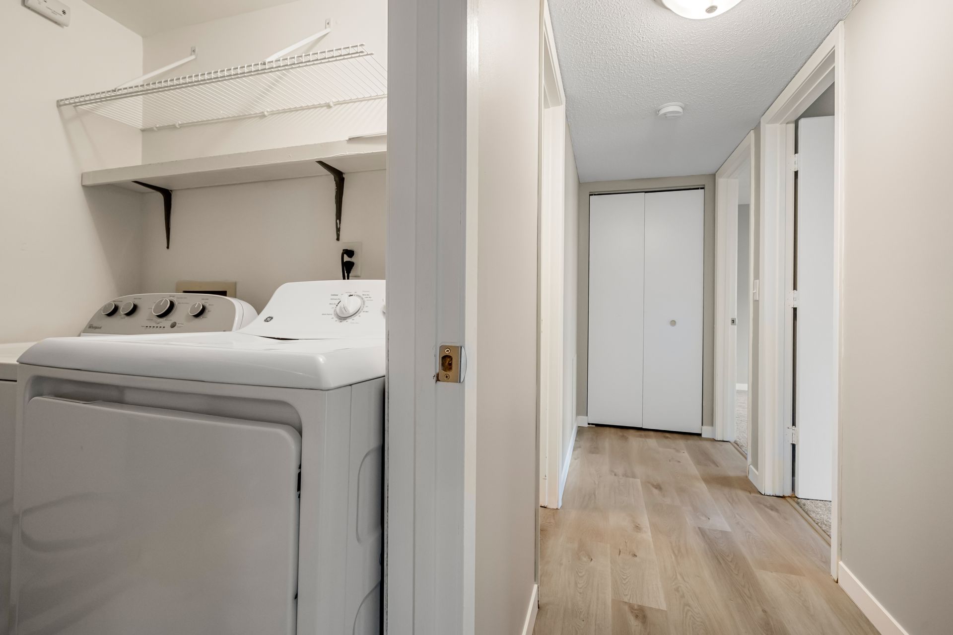 Laundry room and hallway in a bright home, with a washer under a shelf and white doors along wood flooring