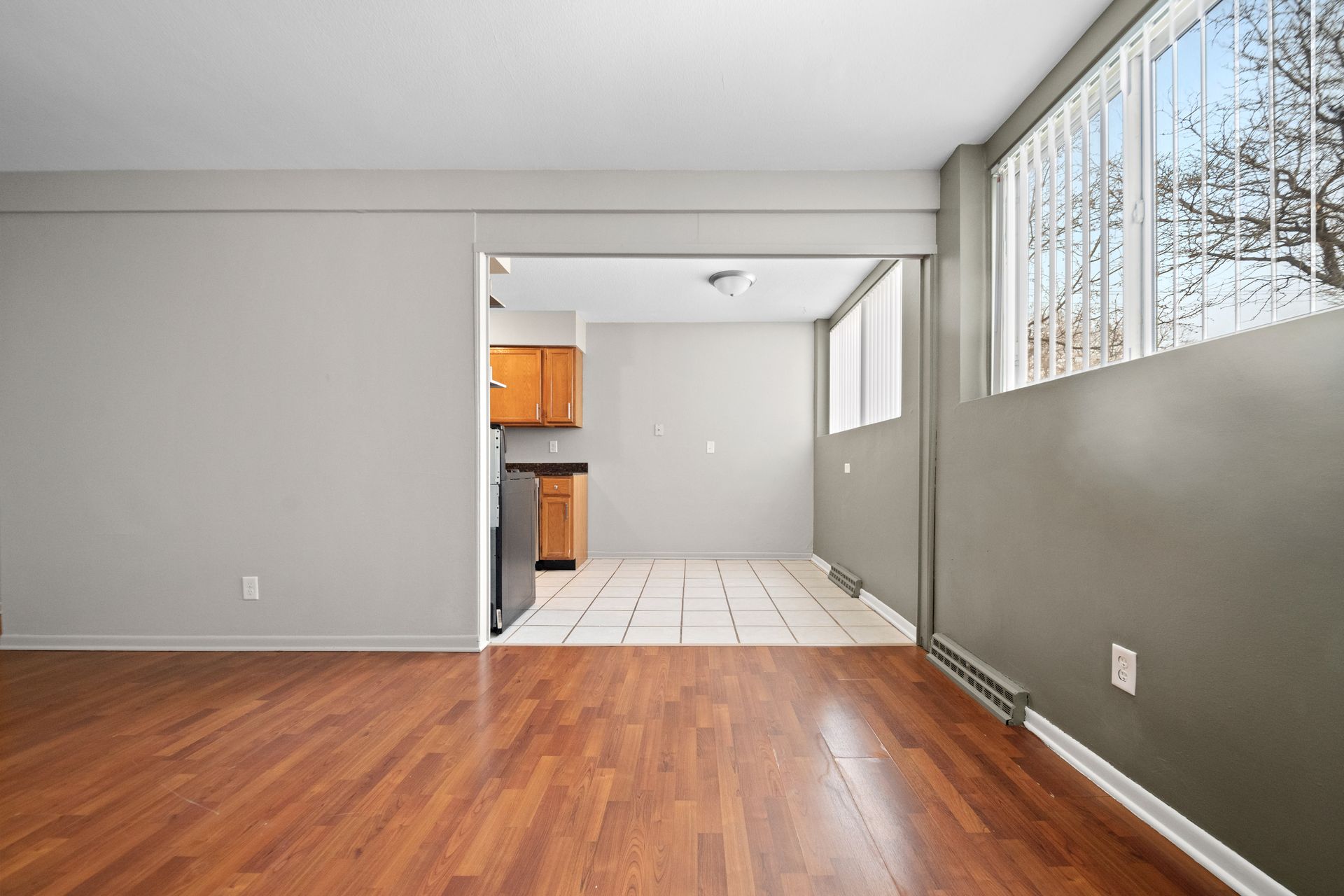 Empty living room with wood floor, gray walls, and a tiled kitchen area with orange cabinets.