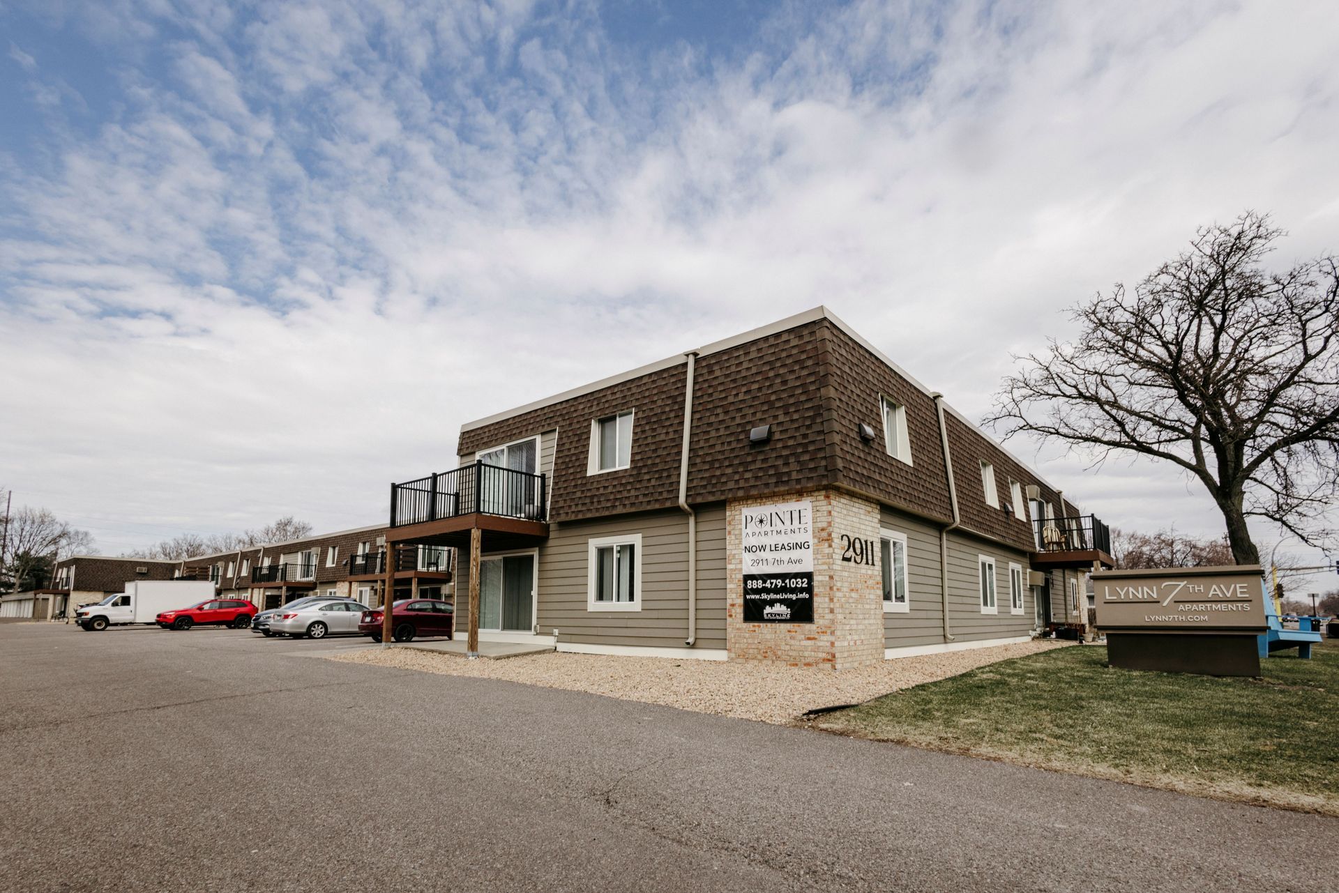 Two-story brown building with parking lot and leafless tree under a cloudy sky