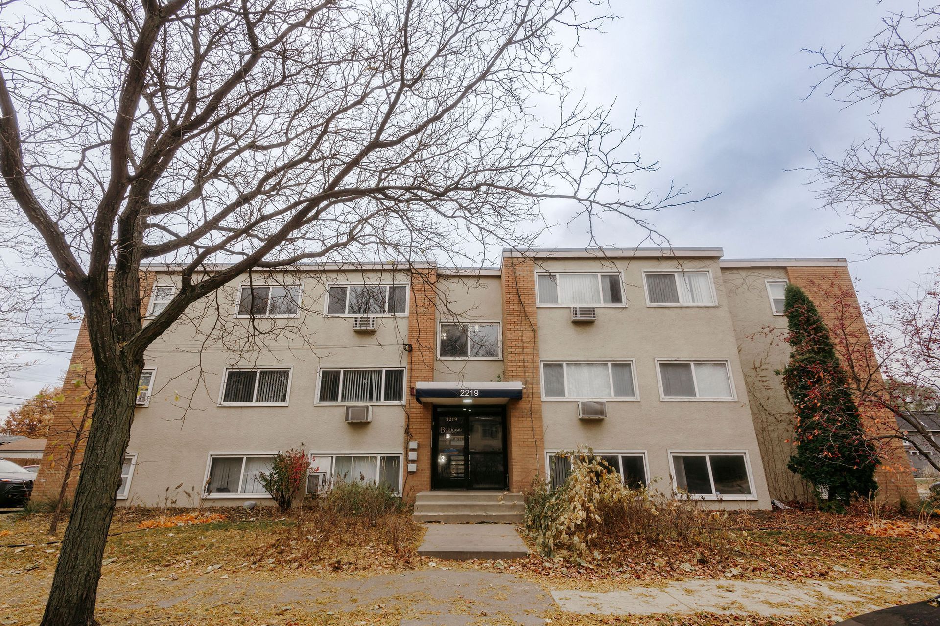 Three-story apartment building with bare trees and fallen leaves in front