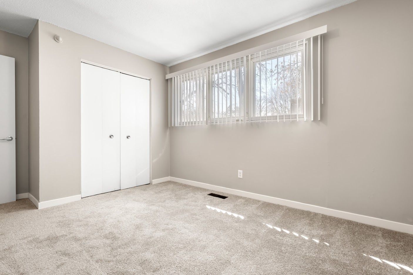 Empty beige bedroom with carpet, closet doors, and two windows with vertical blinds