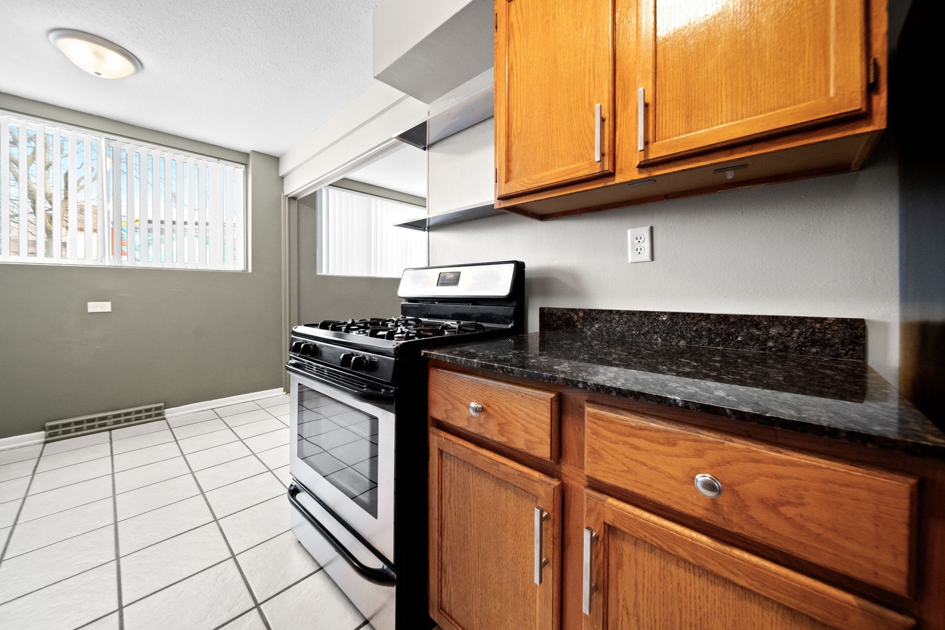 Small kitchen with white tile floor, wood cabinets, granite counter, and a stainless stove by a window.